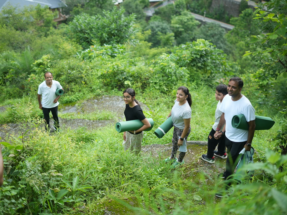 Family carrying yoga mats walking through lush village path near ashram