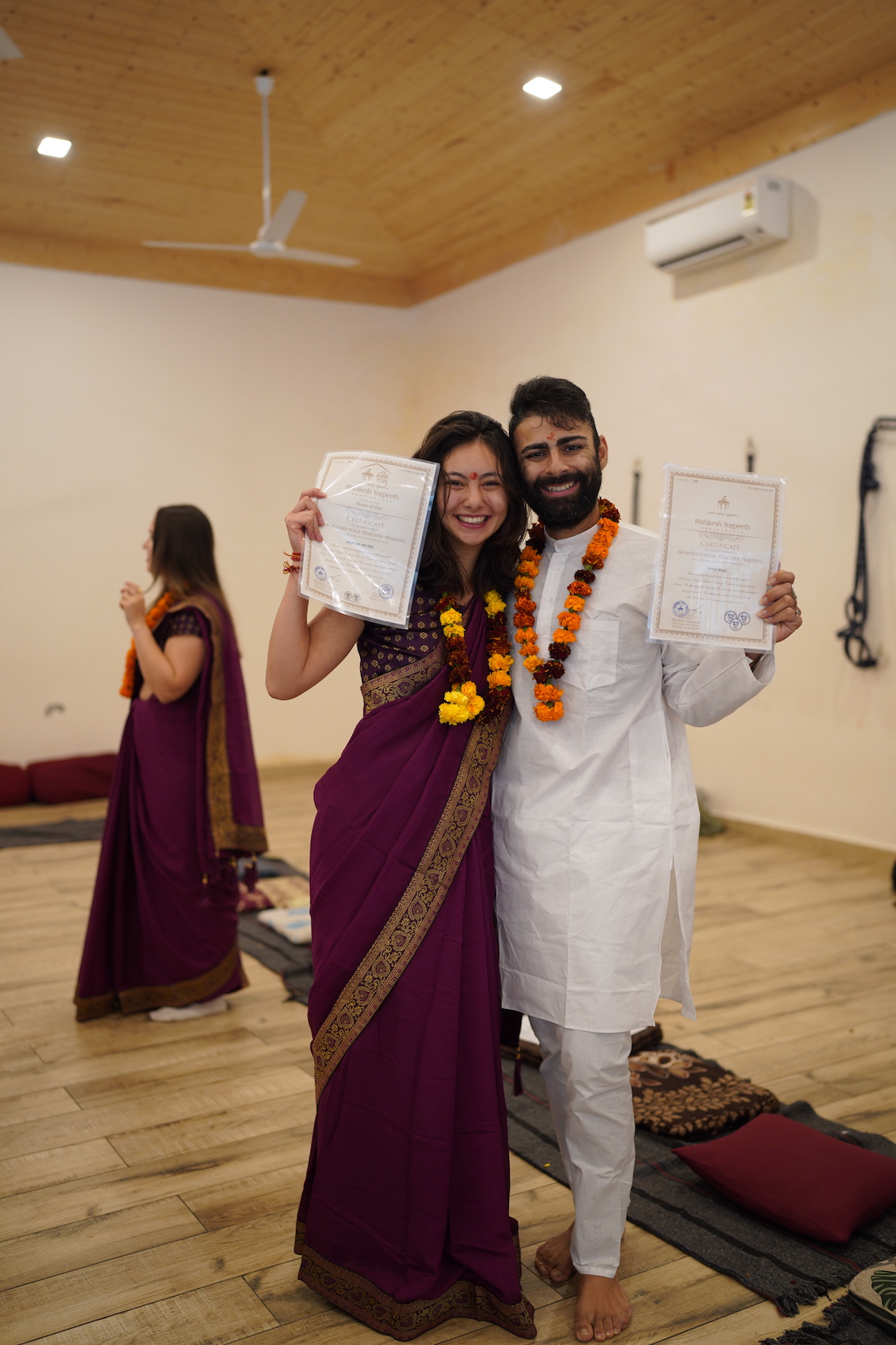 Two graduating students proudly hold up their Rishikesh Yogpeeth Yoga Teacher Training certificates on graduation day
