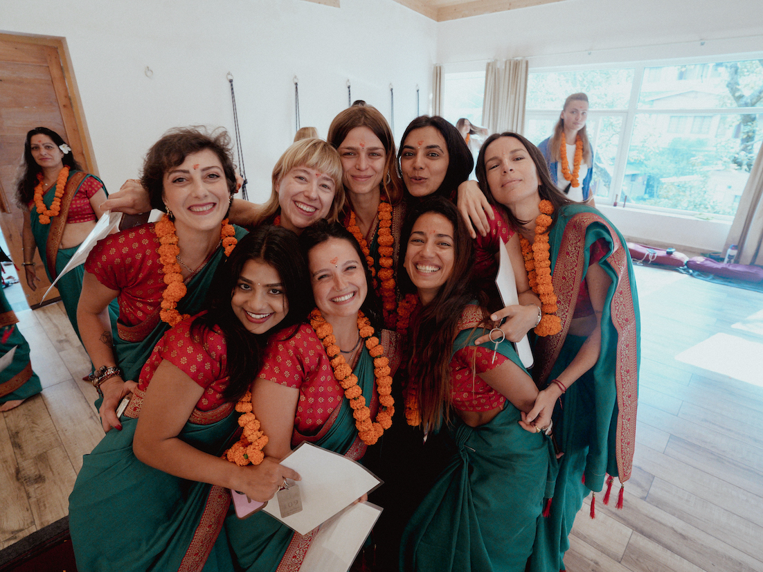 Graduating class of female yoga students embracing in teal saris and marigold garlands on certification day
