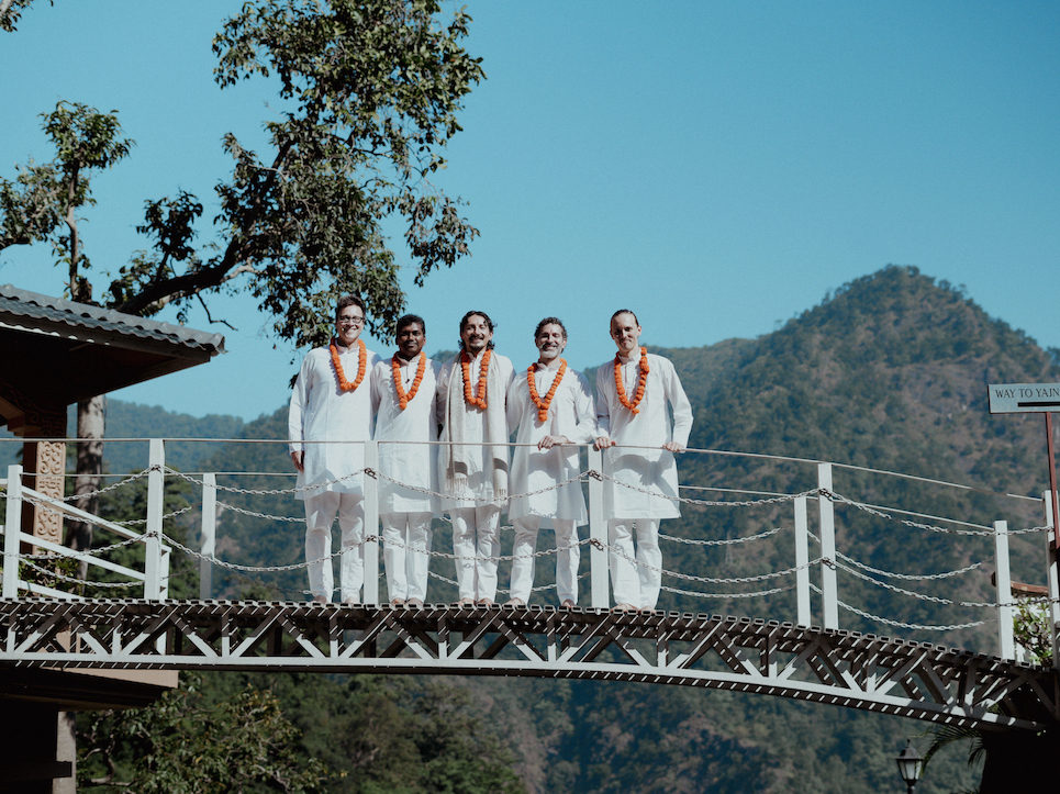 Senior Indian teachers and graduating students standing in white kurtas with marigold garlands on the ashram footbridge