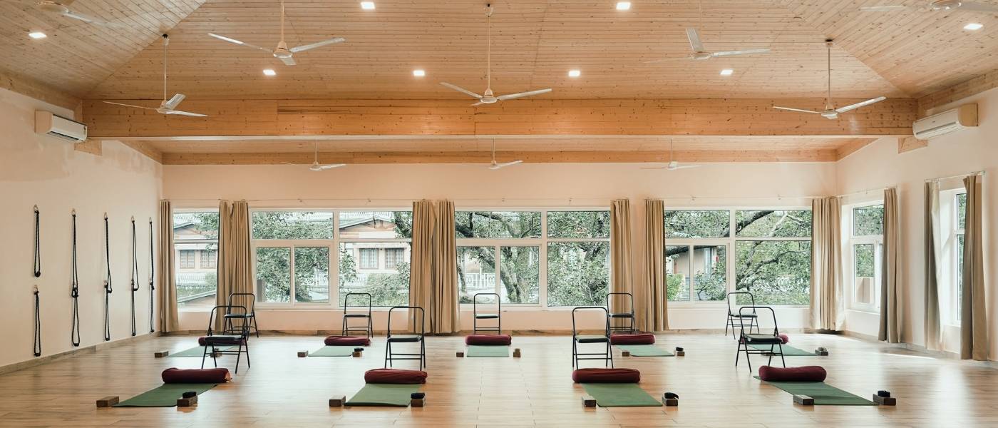 Spacious light-filled yogashala with Iyengar props and green mats at Rishikesh Yogpeeth, yoga teacher training India